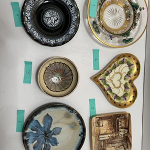 May include: A collection of decorative plates and dishes. Includes a black plate with white floral designs, a clear glass plate with a gold rim and a forest scene, and a heart-shaped dish with floral accents.