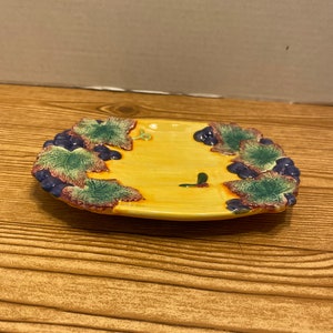 May include: A small, yellow ceramic plate with a grapevine design. The plate has a raised rim and is decorated with green leaves and purple grapes.
