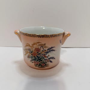 May include: A peach-colored ceramic bowl with two small handles. The bowl is decorated with a floral design in shades of red, blue, and yellow. The rim is trimmed with gold detailing.
