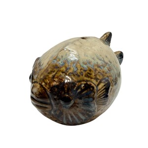 May include: A ceramic pufferfish figurine with a glossy finish. The sculpture features a rounded body with brown, beige, and blue-green hues. Details include fins and facial features, creating a whimsical aquatic theme.
