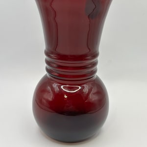 May include: A ruby red glass vase with a bulbous base and a flared top. The vase has a series of horizontal ridges around the middle. The glass is transparent, allowing light to pass through, and the vase is set against a white background.