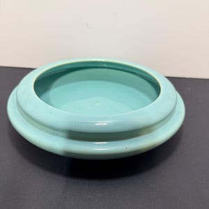 May include: A round, light turquoise ceramic bowl with a wide, flat rim. The bowl has a smooth, glossy finish and a shallow interior. The rim is slightly raised, creating a defined edge. The bowl is likely for decorative or functional use.