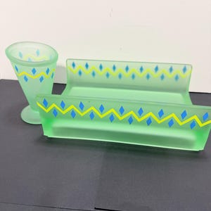 May include: A set of three frosted green glass serving pieces. The set includes a cone-shaped glass, a rectangular tray, and a long, narrow tray. Each piece is decorated with a repeating pattern of blue diamonds and yellow zigzags.