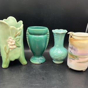May include: Four vintage ceramic vases in different styles and colors. One vase is green with a floral design, another is a green and white vase with a floral design, another is a green vase with a floral design, and the last is a white vase with a landscape scene.