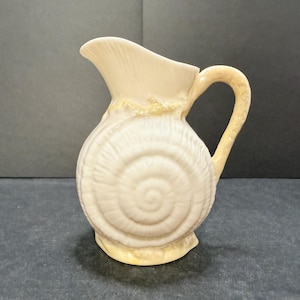 May include: A cream-colored ceramic pitcher shaped like a spiral seashell. The body features a prominent embossed swirl pattern resembling a nautilus shell, with a curved handle and a wide, flared pouring spout at the top rim of the vessel.