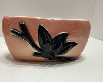 May include: A pink ceramic planter with a black stylized flower design. The planter is oval shaped and has a glossy finish.