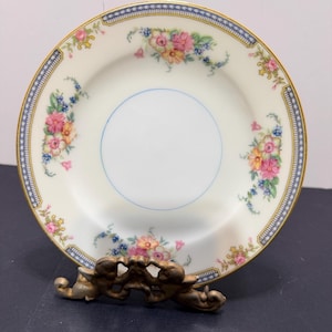 May include: A white porcelain plate with a floral design featuring pink, yellow, and blue flowers. The plate has a gold rim and a blue border with a star pattern. The plate is displayed on a decorative gold-colored stand.