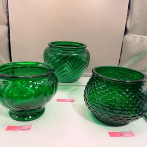 May include: Three green glass bowls with varying textures. One bowl has a swirl pattern, another has a diamond pattern, and the third has a ribbed design. Each bowl has a dark rim and sits on a white surface.