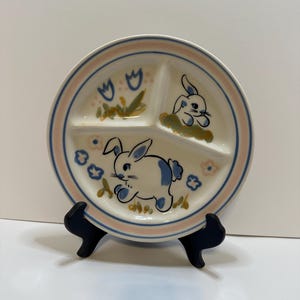 May include: A divided ceramic plate with a pink and blue border. The plate features three sections, each decorated with a cartoon bunny and floral designs. The bunnies have blue spots. The plate is on a black stand.