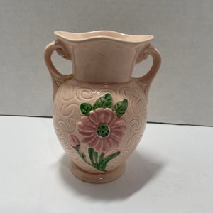 May include: A pink ceramic vase with two handles and a floral design. The vase has a swirl pattern and a pink flower with green leaves.