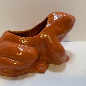 May include: A ceramic frog-shaped planter in a glossy orange-brown glaze. The planter has a wide opening and detailed features, including textured skin and defined frog legs. The decorative piece is suitable for indoor or outdoor use.