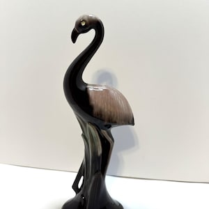 May include: A ceramic flamingo figurine in shades of brown and tan. The bird has a long, curved neck and stands on a pedestal base. The flamingo's feathers are a light tan color, and the beak is dark brown.