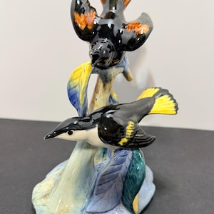 May include: A ceramic figurine featuring two birds perched on a branch. The top bird is black with orange wing and tail accents. The bottom bird is black and white with yellow wing and tail tips. The base is light blue with green and blue leaves.