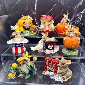 May include: A collection of whimsical mouse figurines. The figurines include mice in a pumpkin, an apple, a magician's hat, and a slot machine. The color palette includes orange, red, white, and brown. The figurines are displayed on clear shelves.