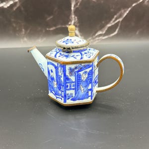 May include: A small, hexagonal teapot with a blue and white design. The teapot features a gold-colored handle and trim, and a lid with a wooden knob. The sides of the teapot depict figures in a landscape.