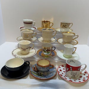May include: A collection of various teacups and saucers. The teacups are in different colors and designs, including gold, red, black, and floral patterns. Some cups have matching saucers, while others have unique pairings. The arrangement suggests a vintage or eclectic aesthetic.