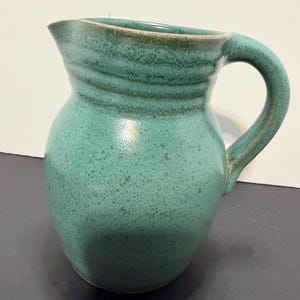May include: A handcrafted, turquoise ceramic pitcher with a rounded body and a curved handle. The pitcher has a spout for easy pouring and a textured surface. The rim and handle have a darker, earthy tone.