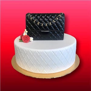 May include: A white cake with a quilted design and a black quilted handbag on top. The cake is decorated with red and white flowers.