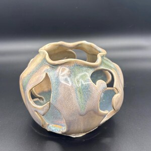 May include: A round ceramic vessel featuring a wavy, open-cut rim and large, irregular cutouts along the sides. The surface has a glossy glaze with blended tones of beige, soft blue, and sage green, creating a smooth, reflective finish throughout.