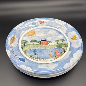 May include: A round ceramic plate featuring a central illustration of a pond with two figures fishing, a red house, and a yellow sun. The border is decorated with a blue sky pattern, white clouds, a yellow kite, and small birds in flight.