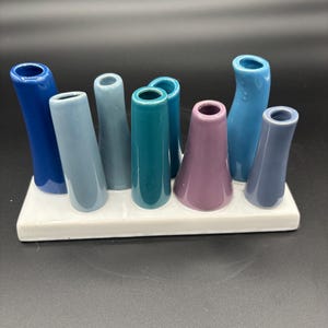 May include: A multi-tube ceramic flower vase featuring eight individual stems of varying heights and shades, including dark blue, light blue, teal, and purple, all attached to a single rectangular white base with a smooth, glossy finish.