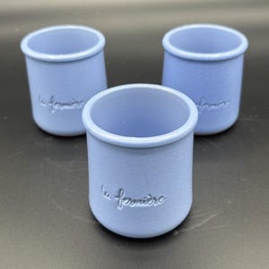 May include: Three light blue ceramic pots with a smooth, matte finish. Each pot features a rounded cylindrical shape with a slightly flared rim and the embossed text la fermière on the front side. The pots are arranged in a triangular formation.