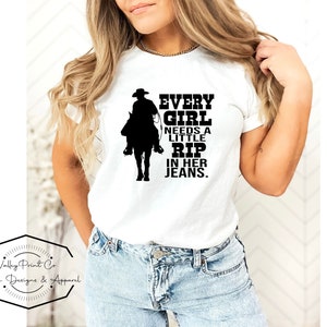 May include: A white short-sleeved t-shirt featuring a black graphic of a cowboy on horseback. The text printed in black reads: EVERY GIRL NEEDS A LITTLE RIP IN HER JEANS. The shirt is paired with light blue denim jeans with a visible metal button.