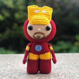 Puede incluir: Un juguete de peluche tejido a crochet de Iron Man. El juguete es rojo y amarillo con un círculo azul en el pecho. El juguete tiene una cara tejida a crochet con ojos negros y una barba marrón.