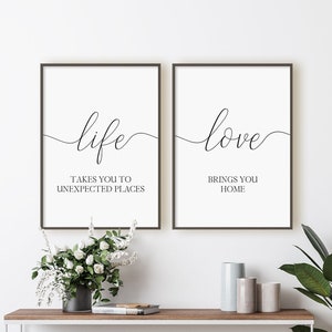 Puede incluir: Dos impresiones enmarcadas con caligrafía negra sobre fondo blanco. La impresión de la izquierda dice "life takes you to unexpected places" y la impresión de la derecha dice "love brings you home". Las impresiones están colgadas en una pared blanca sobre una mesa consola de madera con un marco de metal blanco. Hay flores blancas en un jarrón sobre la mesa y una planta verde en maceta en una cesta tejida en el suelo.