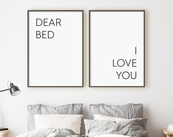 May include: Two framed prints with black text on a white background. The left print says "Dear Bed" and the right print says "I Love You". The prints are hanging on a white wall above a bed with gray bedding.