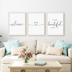 Op de afbeelding: Drie ingelijste prints met witte passe-partouts en zwarte kalligrafie. De prints tonen "welcome", een hart symbool en "to our beautiful chaos". De prints hangen aan een witte muur boven een witte bank met kussens.