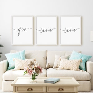 Könnte beinhalten: Drei weiße gerahmte Drucke mit schwarzer Kalligraphie auf weißem Hintergrund. Die Drucke lauten "que sera sera" in einer kursivschriftlichen Schriftart. Die Drucke hängen an einer weißen Wand über einem weißen Sofa mit Kissen.