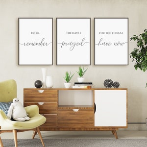 May include: Three framed wall art prints with black frames and white backgrounds. Each print has a different inspirational quote in black script: "I still remember", "The days I prayed", and "For the things I have now". The prints are displayed above a wooden cabinet.