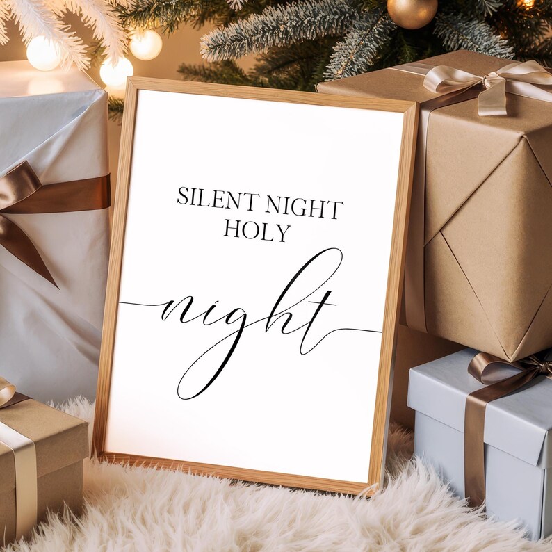 May include: A framed print with the words "SILENT NIGHT HOLY night" in black script. The wooden frame is light brown. The print is surrounded by wrapped gifts with brown and blue paper, and a Christmas tree in the background.