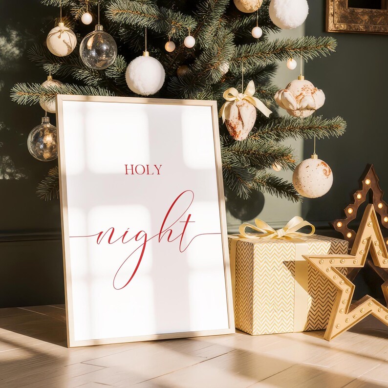 May include: A framed print with the words "HOLY night" in red script. The print is propped up in front of a decorated Christmas tree with ornaments and a wrapped gift box. A wooden star with lights is also visible.