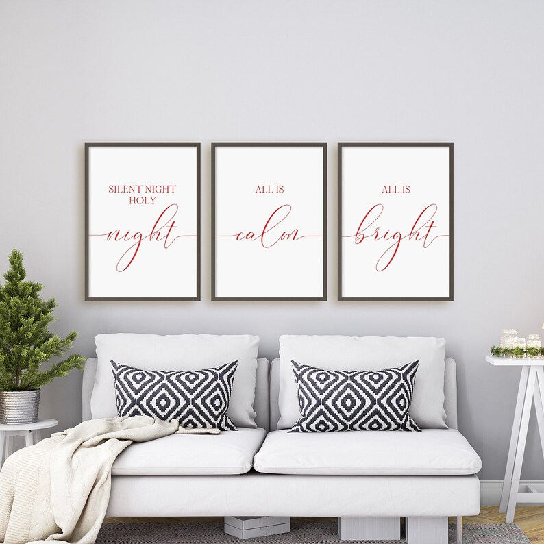 May include: Three framed prints with holiday-themed text in red script. The prints are displayed above a white couch with patterned pillows. A small Christmas tree and side table are visible on the left.