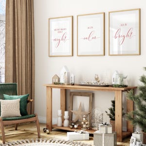May include: Three framed prints with gold trim, each with red script: "Silent Night Holy Night", "All is Calm", and "All is Bright". A wooden console table displays decorative items, including houses, candles, and a star, with wrapped gifts below.