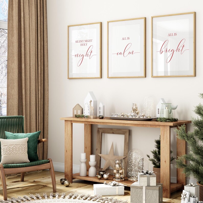 May include: Three framed prints with gold trim, each with red script: "Silent Night Holy Night", "All is Calm", and "All is Bright". A wooden console table displays decorative items, including houses, candles, and a star, with wrapped gifts below.