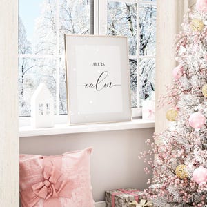 May include: A framed print with the words "ALL IS calm" in black script sits on a windowsill. A white house-shaped candle holder and a pink pillow with a bow are in the foreground. A Christmas tree with pink and gold ornaments is on the right.