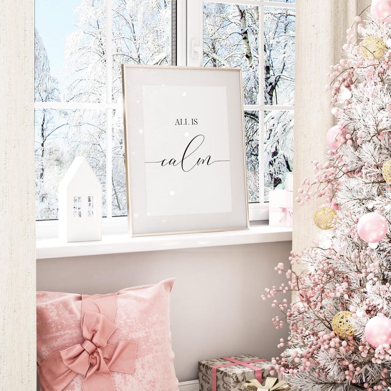 May include: A framed print with the words "ALL IS calm" in black script sits on a windowsill. A white house-shaped candle holder and a pink pillow with a bow are in the foreground. A Christmas tree with pink and gold ornaments is on the right.