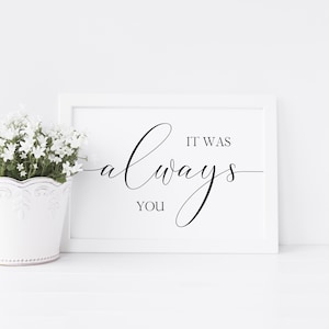 May include: A white framed print with the words "It Was Always You" in elegant black script. A white flower pot with white flowers sits to the left of the print. The background is white.