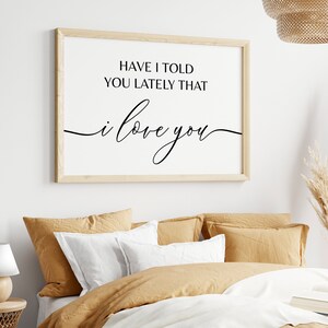May include: A framed print with a white background and black text that reads "Have I told you lately that I love you". The print is hanging on a white wall above a bed with tan bedding.