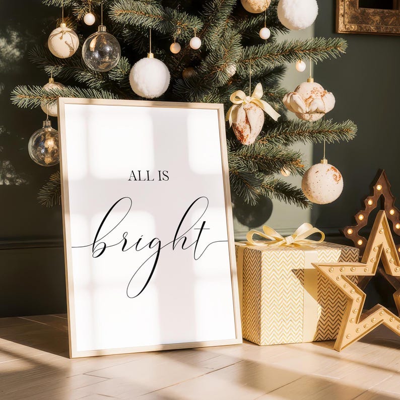 May include: A framed print with the words "ALL IS bright" in elegant script. The print is propped up in front of a decorated Christmas tree with ornaments and a wrapped gift box with a bow. A star-shaped decoration is also visible.
