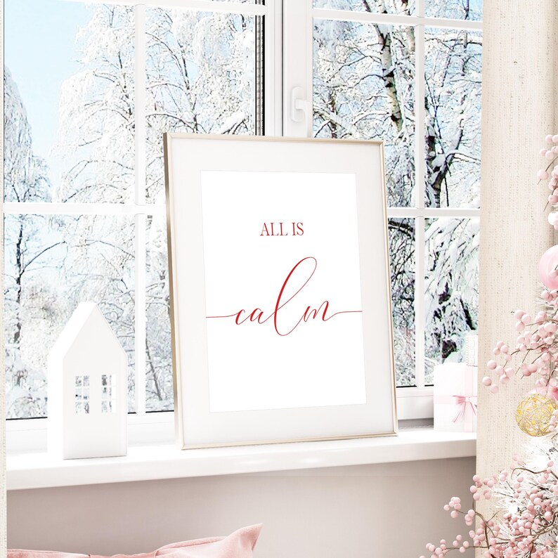 May include: A framed print with the words "ALL IS calm" in red script. The print is displayed on a windowsill with a snowy winter scene visible through the window. A white decorative house and pink decorations are also present.