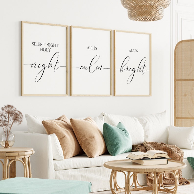 May include: Three framed wall art prints with the words "Silent Night Holy Night", "All is Calm", and "All is Bright" in elegant script. The prints are in light wood frames, hanging above a white sofa with tan and teal pillows. A round coffee table and side table are in the foreground.