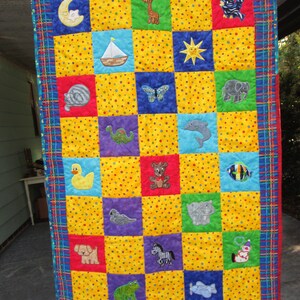 Peut inclure: Une courtepointe pour bébé jaune et bleue avec une variété de motifs d'animaux et d'objets brodés. La courtepointe est faite de carrés avec un fond jaune et des pois blancs. La bordure est un plaid rouge, bleu et vert.