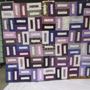 May include: A purple and white quilt with a geometric pattern. The quilt is made of various shades of purple and white fabric, and the pattern is created by using different shapes and sizes of fabric squares. The quilt is hanging on a wall, and the date 04/15/2018 07:57 is visible in the bottom right corner.