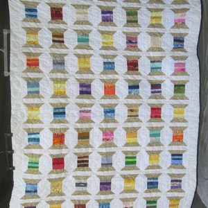 May include: A white quilt with a pattern of brown spools of thread. Each spool is made of different colored fabric strips. The quilt is made up of 49 spools.