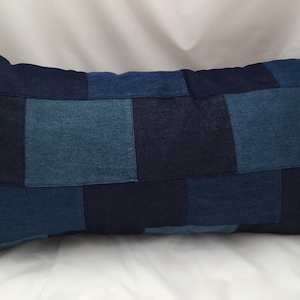 May include: A long rectangular cushion made of denim fabric. The cushion is made of different shades of blue denim fabric sewn together in a patchwork pattern.