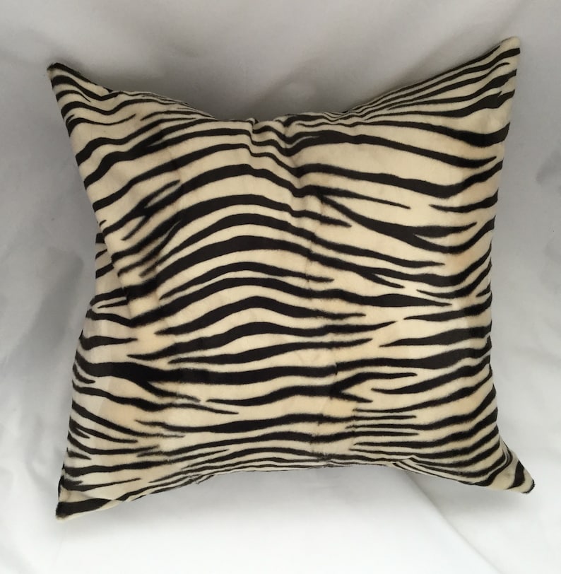May include: Square decorative pillow with a zebra stripe pattern. The pillow features a cream-colored background with bold black stripes, creating a striking contrast. The pillow is a soft, plush home decor item.